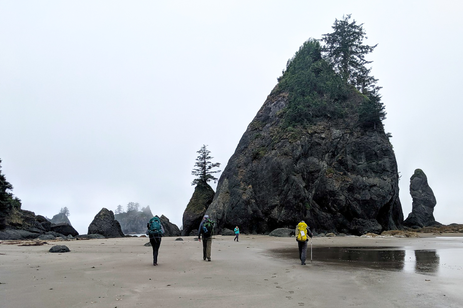 Three hikers walk along a moody beach, with a big rock jutting up in front of them.