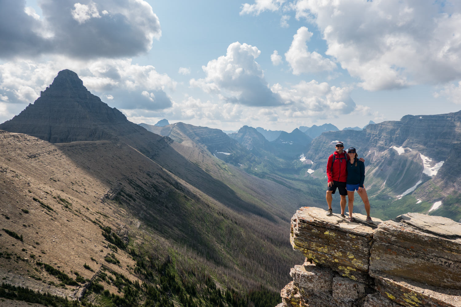 two hikers on a rocky ledge overlooking a deep valley in glacier national park.