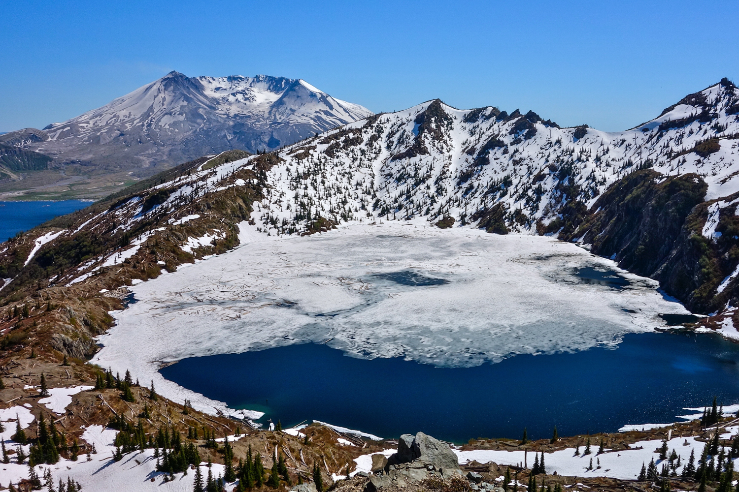An alpine lake is partially frozen over, with snow-capped peaks flaking three sides.
