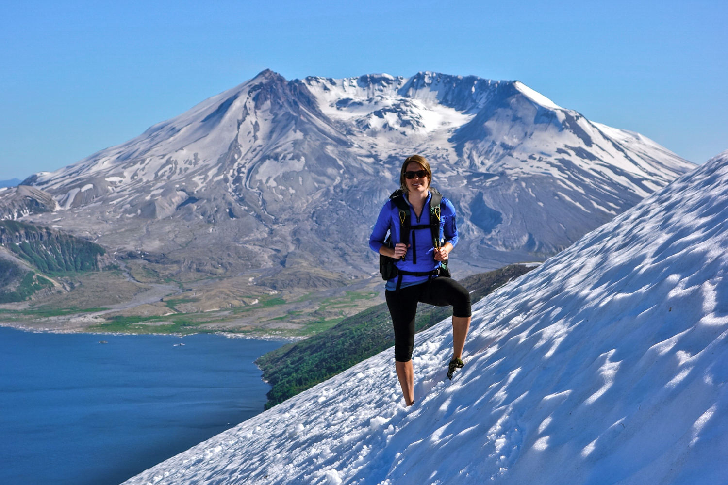 A female hiker smiles at the camera while standing on a steep, snowy slope with views of an alpine lake and snow-capped peak in the background.