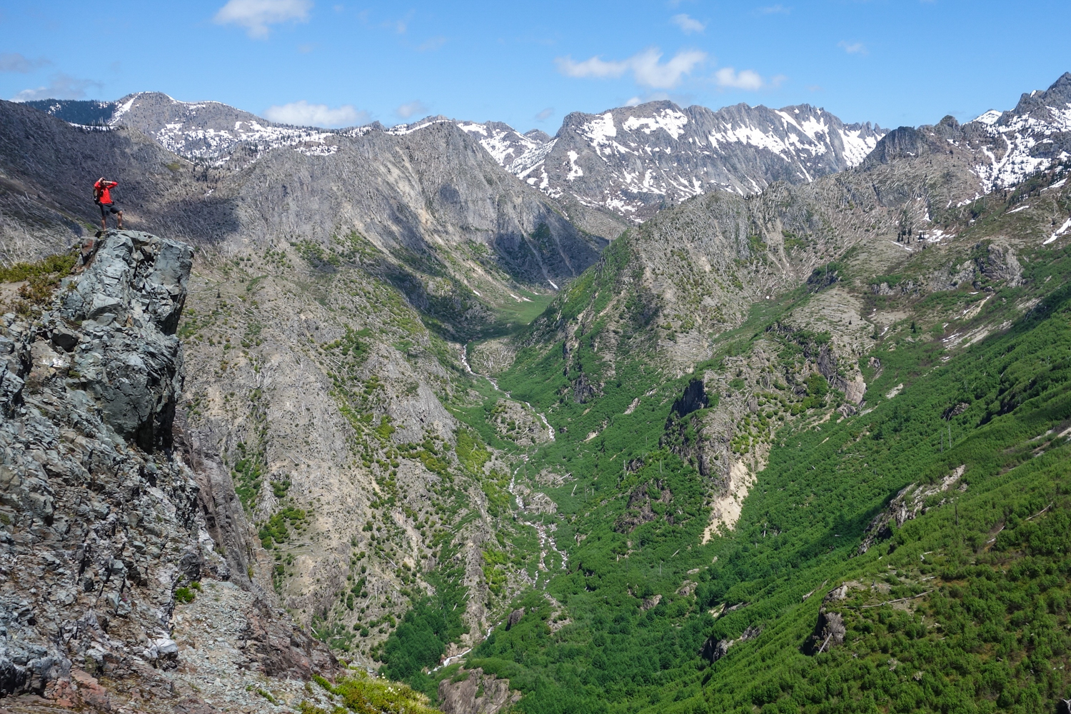 A hiker stands on a rocky outcropping with big views of surrounding mountains all around him.