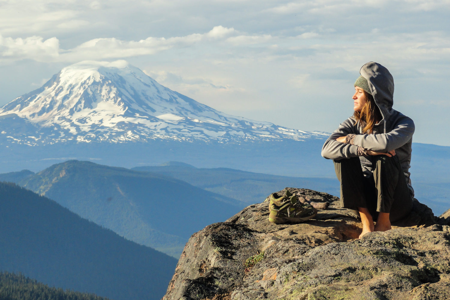 A woman sits with her eyes closed on a rock with a view of towering Mt. Rainier in the distance.