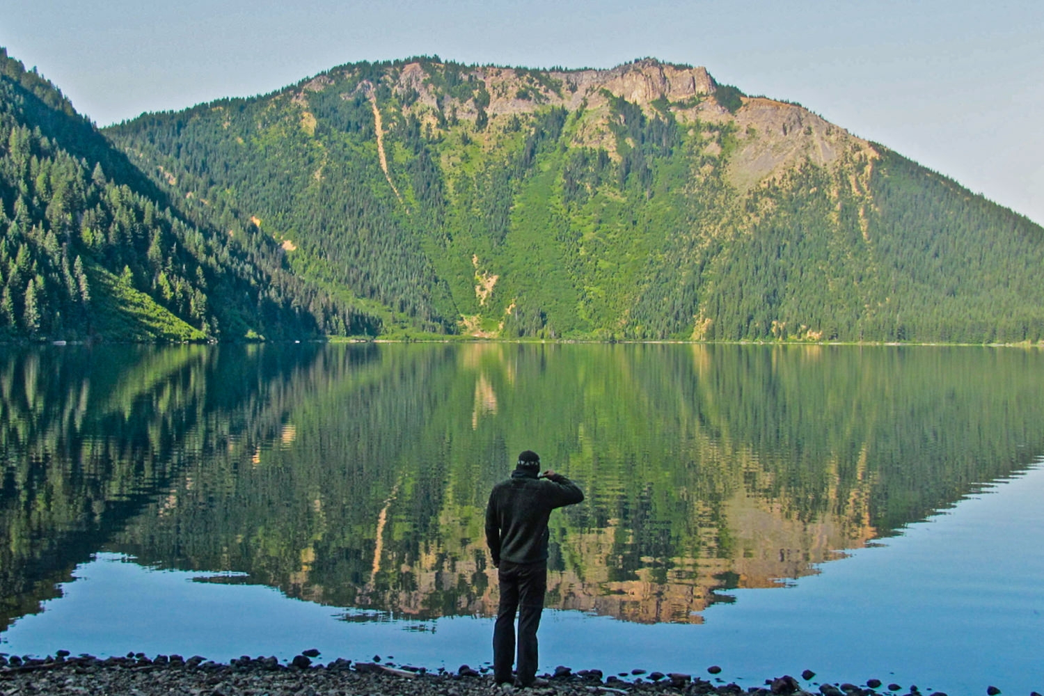 A man stands at the edge of a lake with a mountain at the other side. The mountain is reflected perfectly in the still water.
