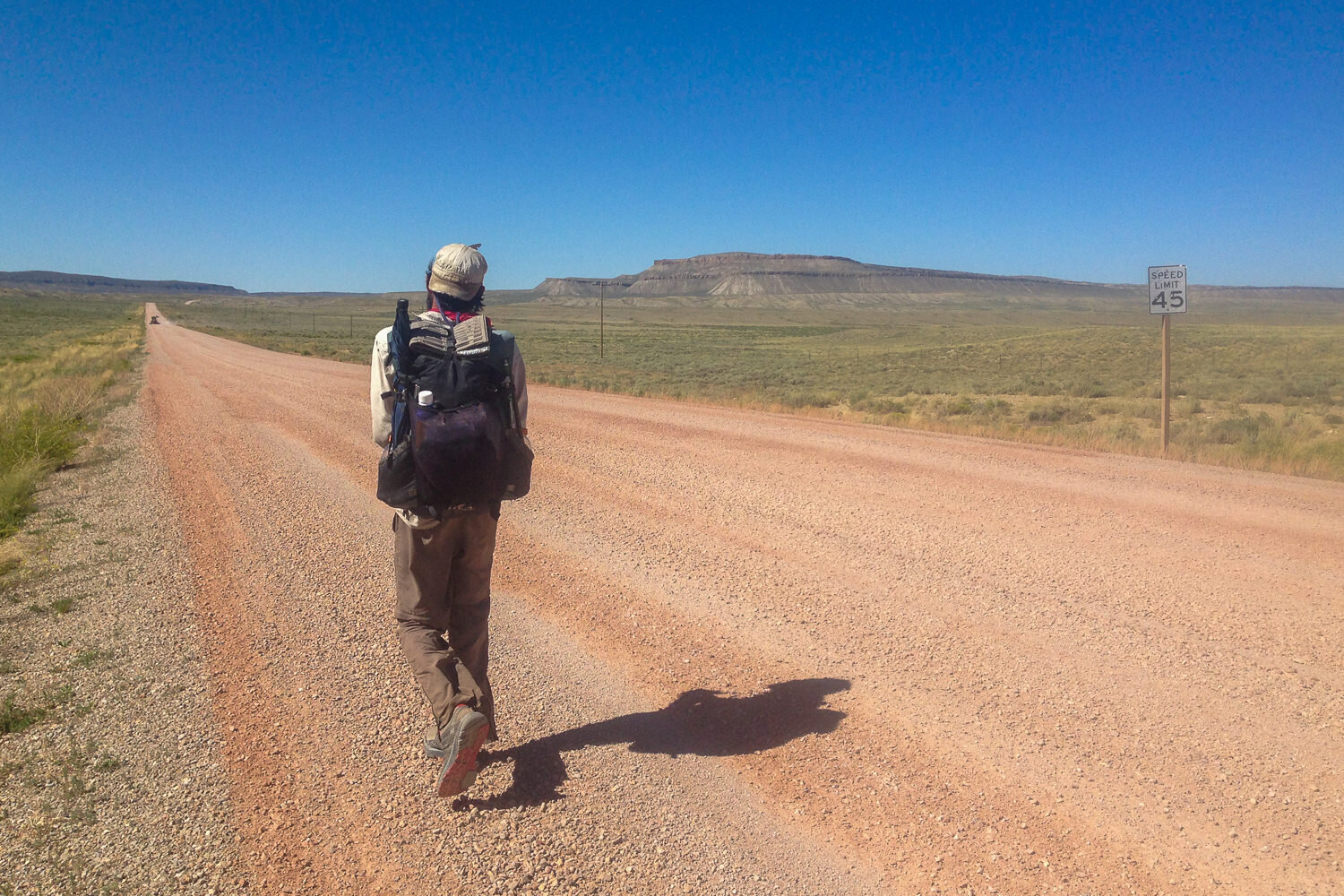 CDT thru-hiker walking along a straight dirt road in the sun with a cloudless blue sky.