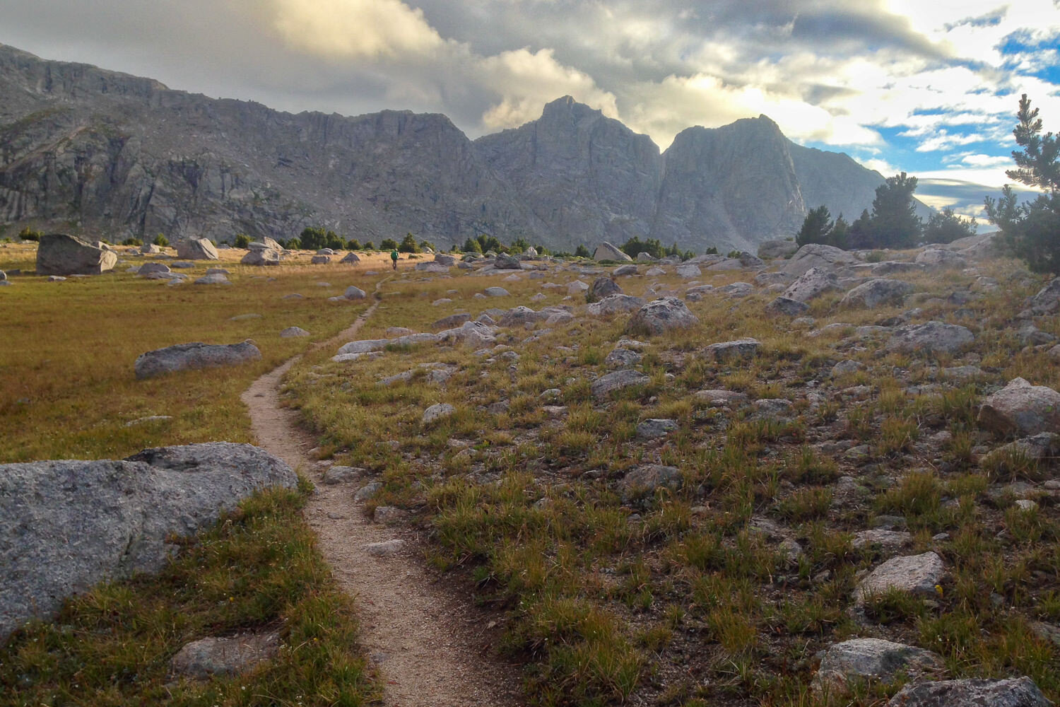 CDT trail meandering through a boulder-filled meadow with mountains in the background.