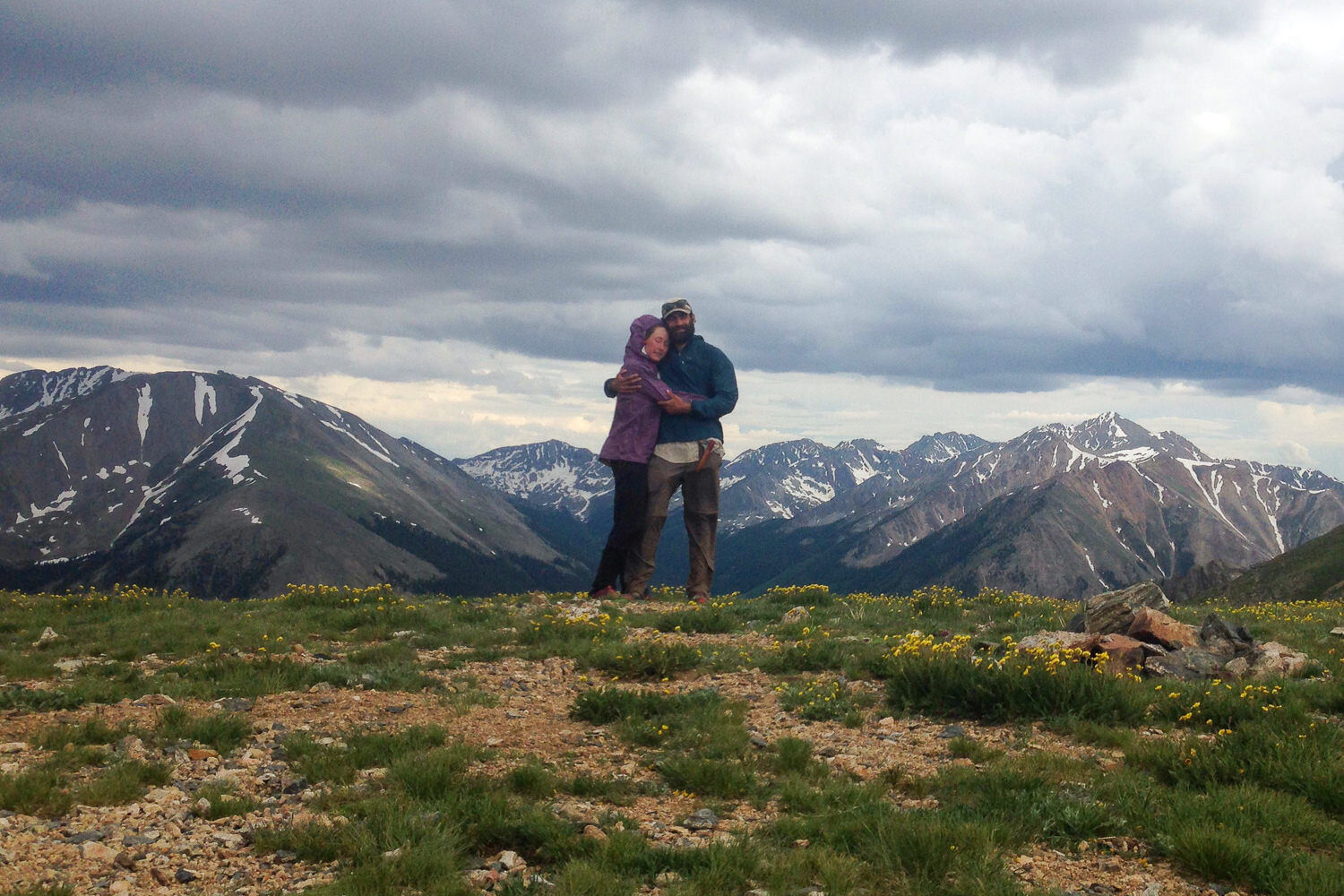Two thru-hikers stop for a hug and picture on top of a beautiful mountain view on a cloudy day.