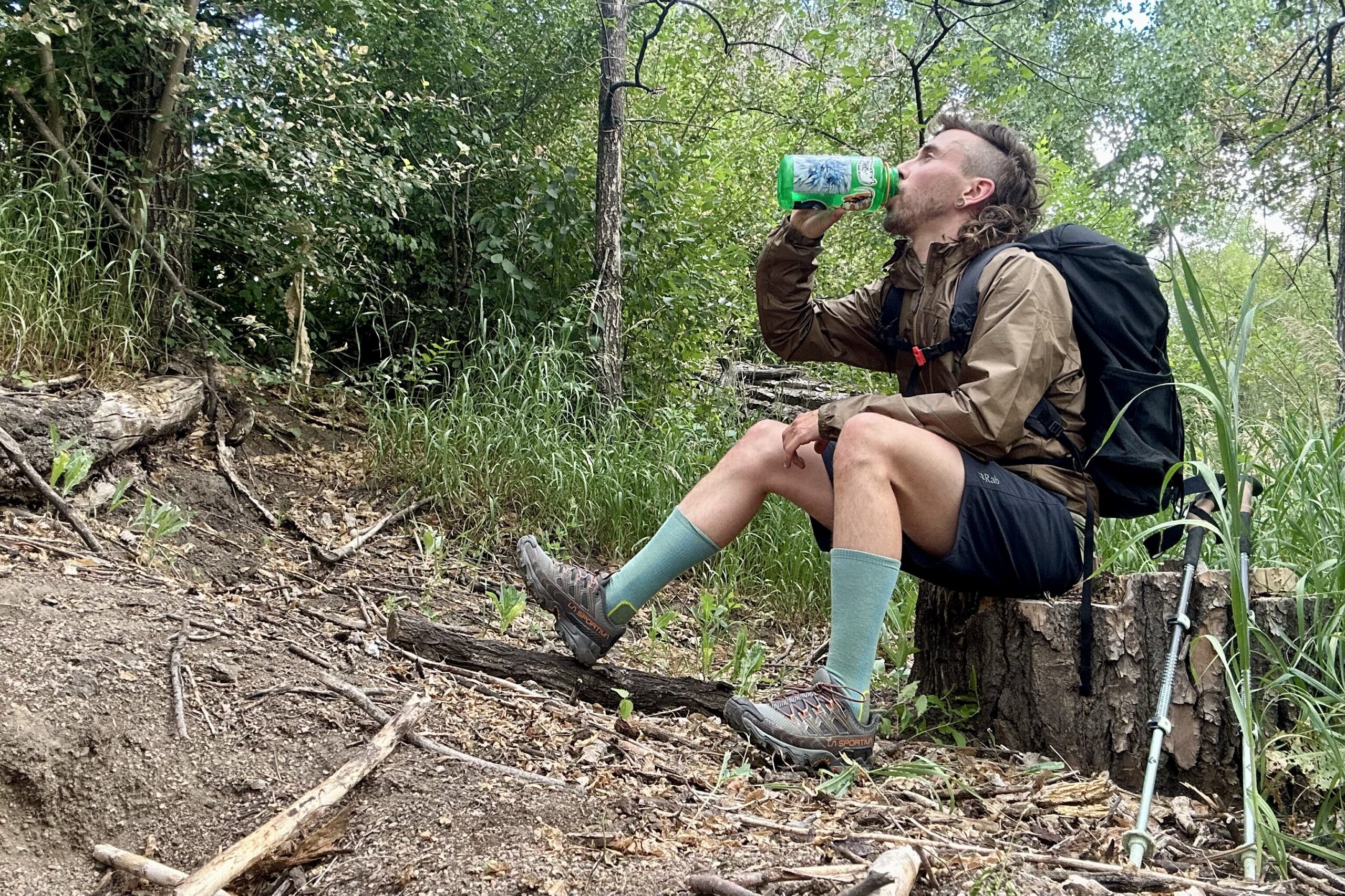 A hiker sits on a tree stump drinking water in a forest setting.