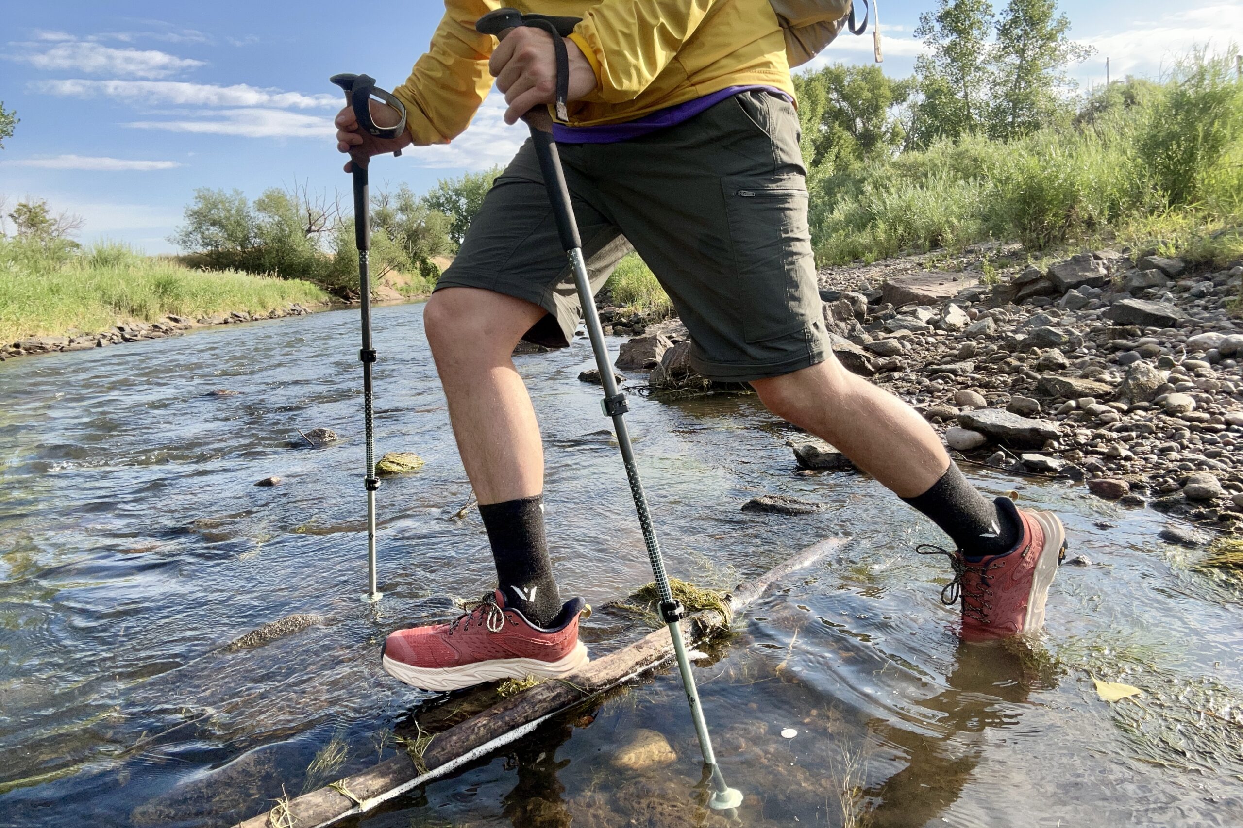 A male hiker walks across a river in a wilderness area using poles.