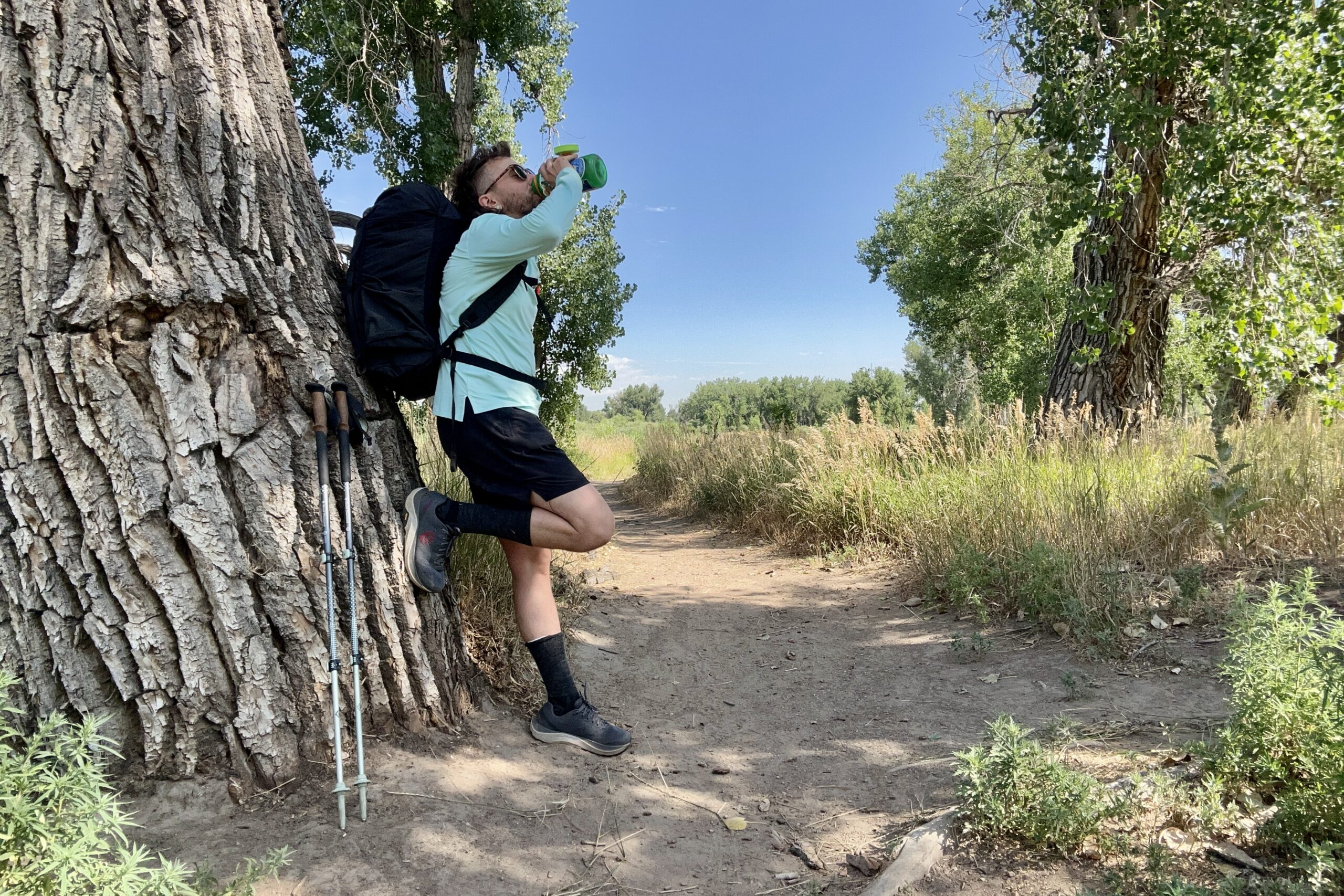 A hiker leans against a tree to stop and drink water on a trail in a sun hoodie under a blue sky.