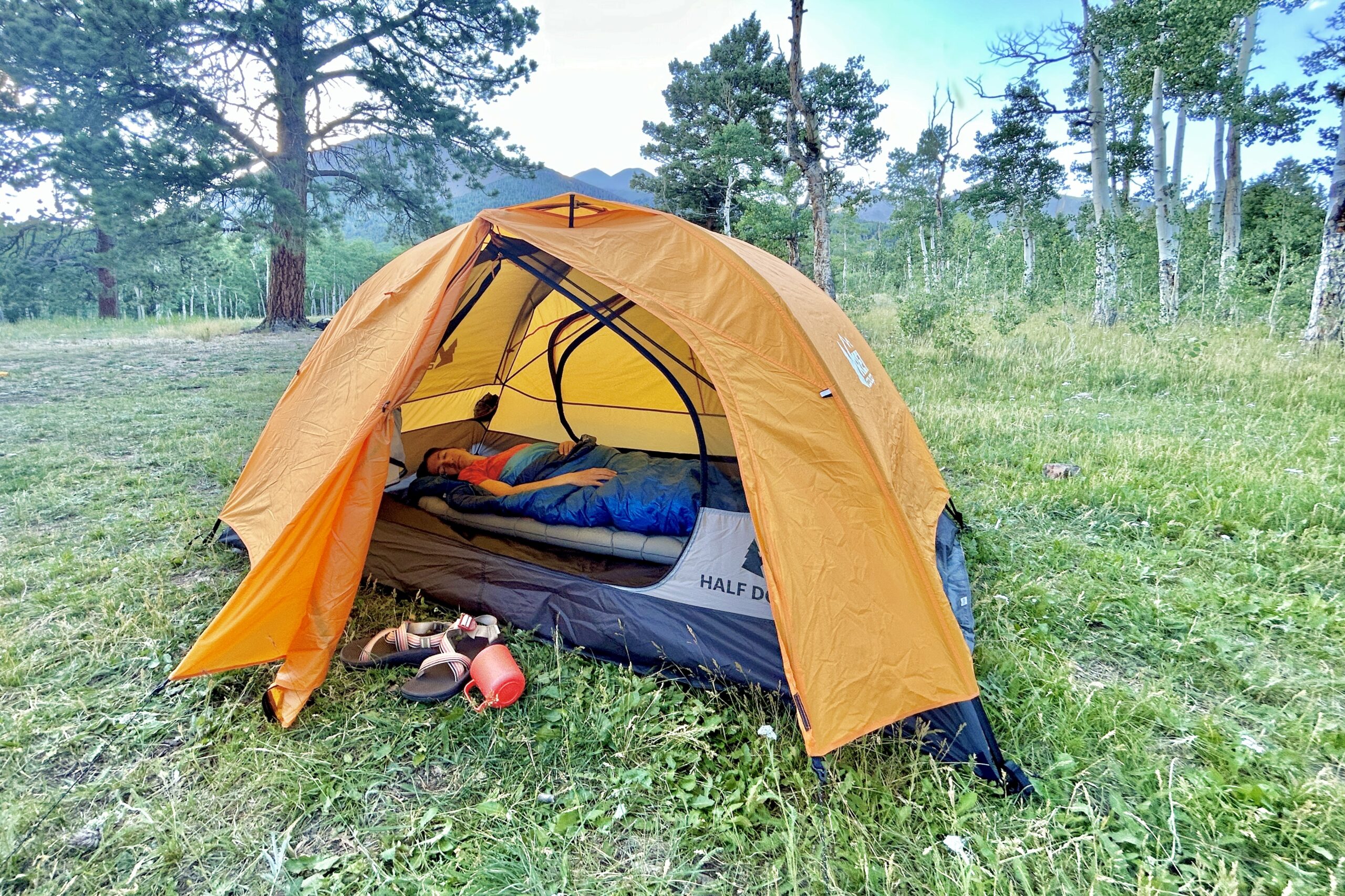A woman in a sleeping bag rests in an orange tent in the woods with mountains in the background.