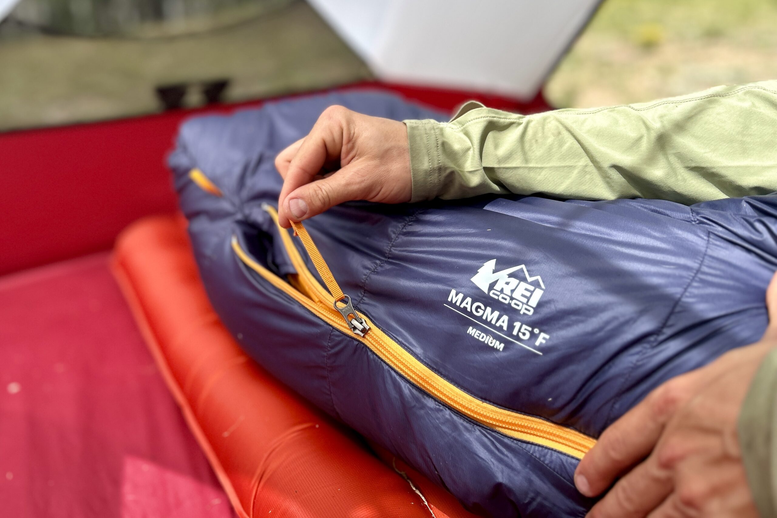 A person zips up a sleeping bag inside a tent. Only their hands and the footbox of the sleeping bag are visible.