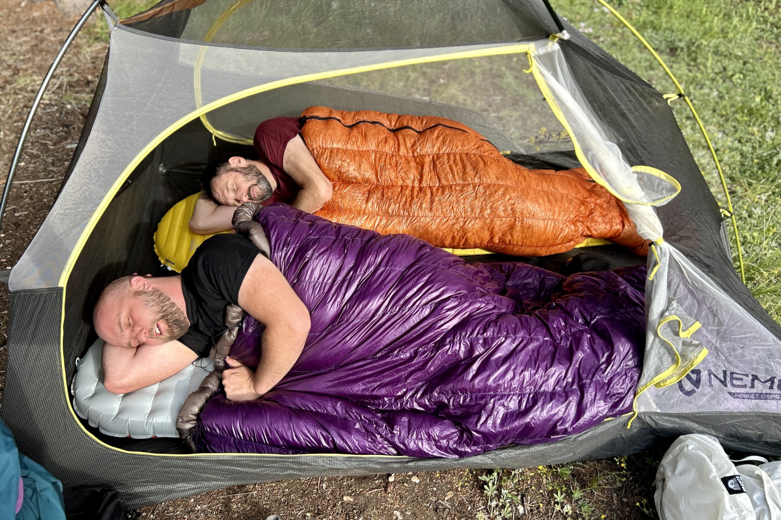 Two men rest inside of sleeping bags, on their own pads, next to each other in a tent on a sunny morning.