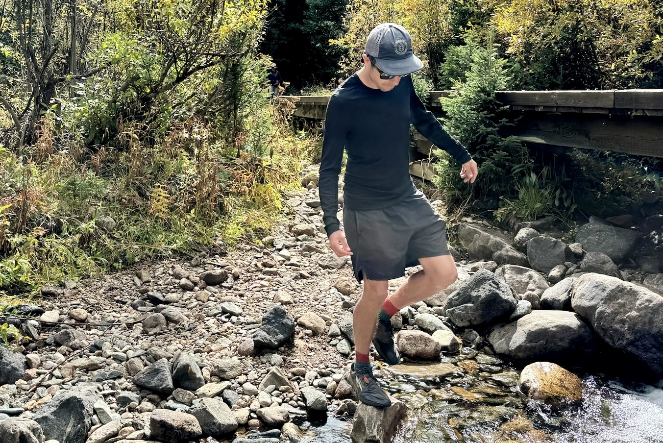 A hiker crossed a stream on rocks while wearing the Icebreaker Merino Oasis 200 base layer. A bridge and trees are in the background.
