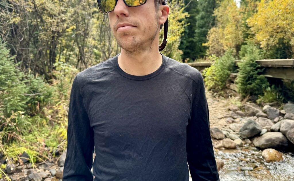 A hiker stands in the woods wearing the Icebreaker Oasis 200 base layer. Trees, a bridge, and a creek are in the background.