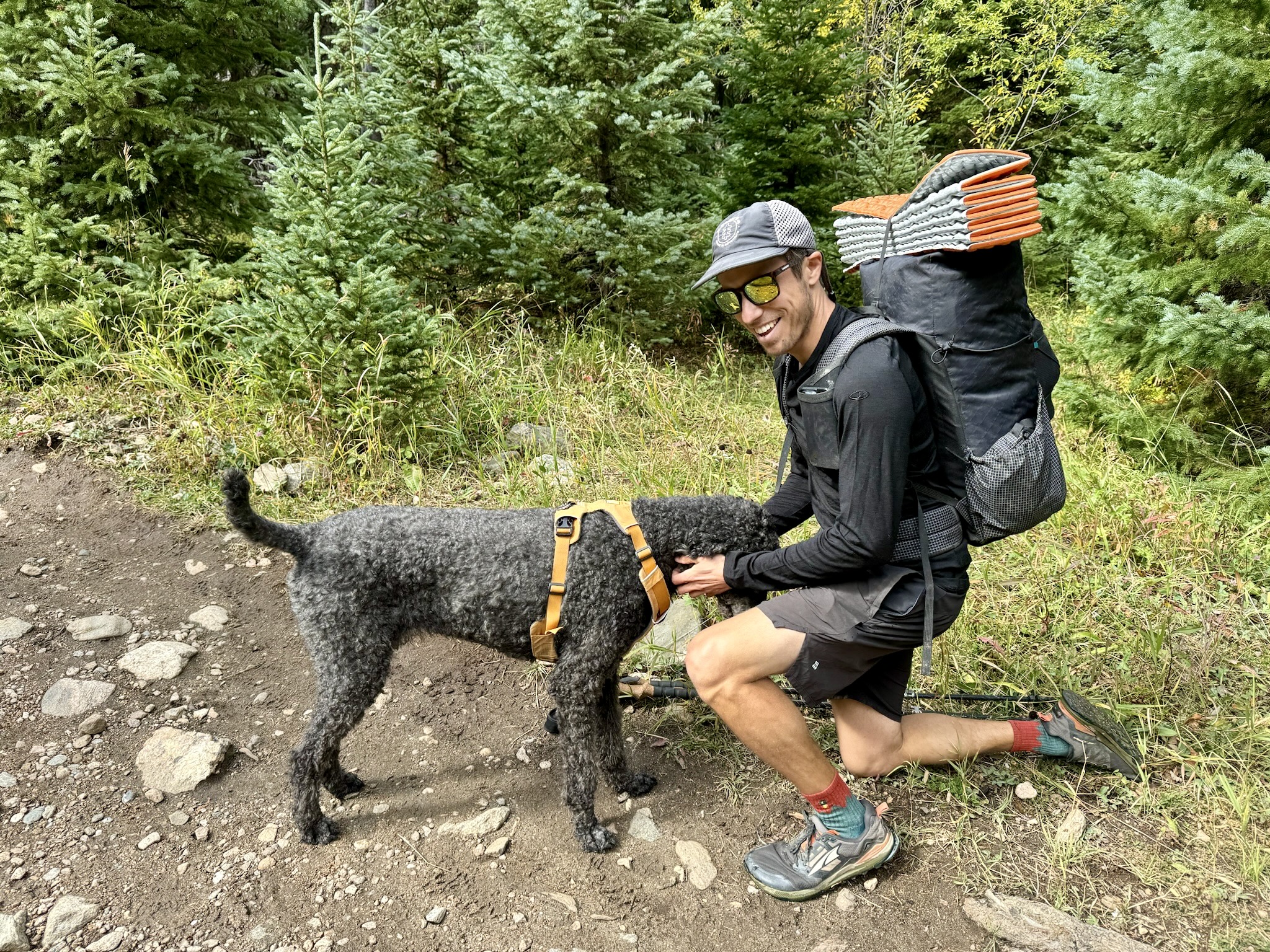 A hiker kneels to pet a dog on a trail while wearing a pack and the Icebreaker Oasis 200 base layer. Trees are in the background.