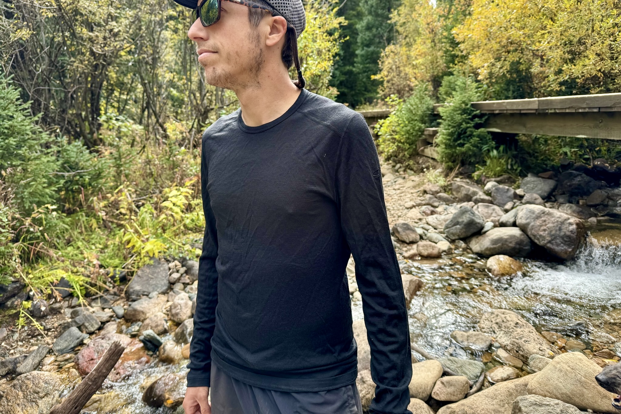 A hiker stands next to a stream wearing the Icebreaker Oasis 200 base layer. Trees and a bridge are in the background.
