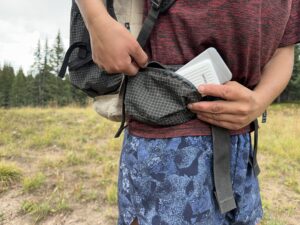 Close up of a backpack side pocket with one hand placing the Zendure and a cell phone into the pocket and the other holding the zipper. There are trees in the background.