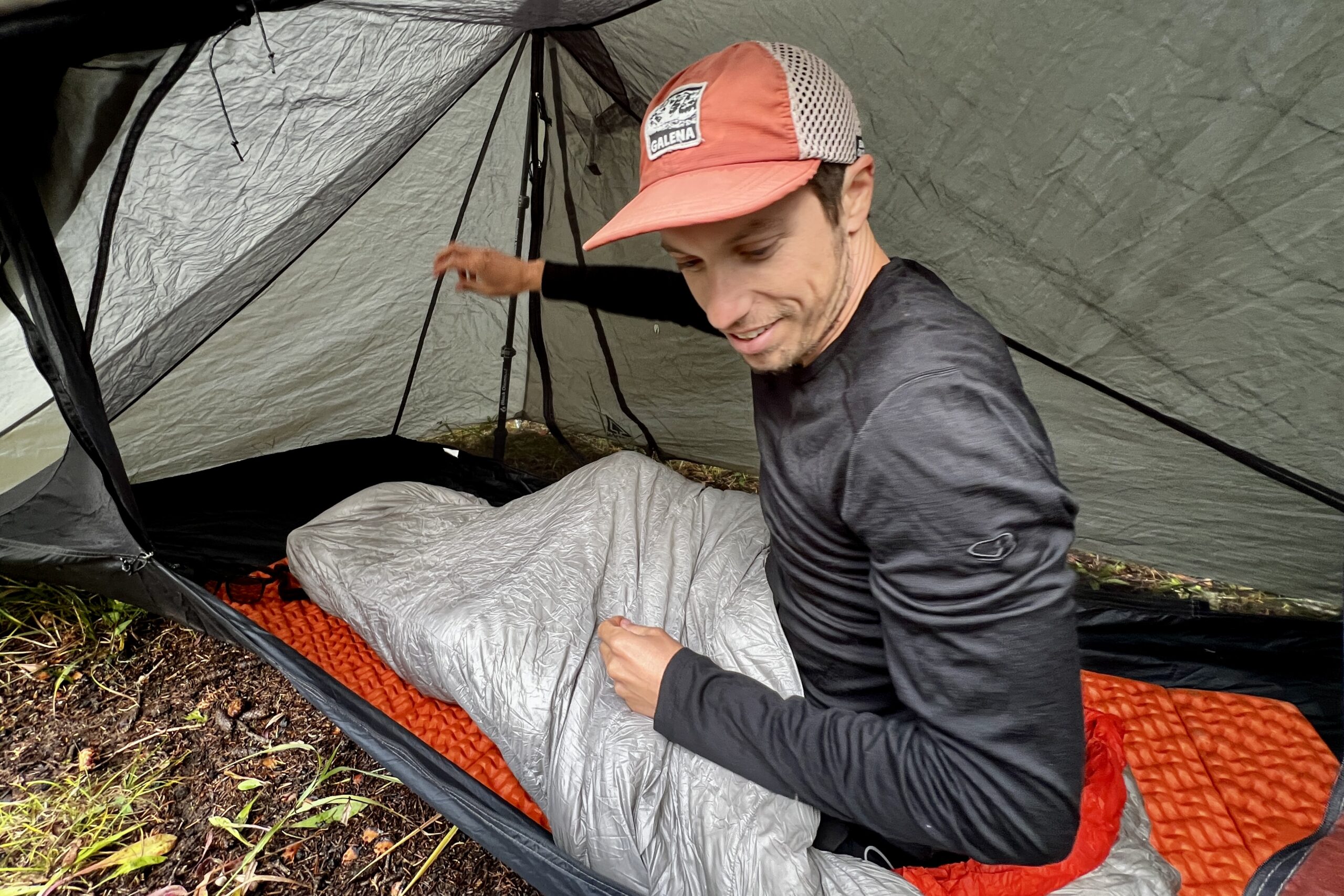A hiker sits in his sleeping bag in a tent while wearing the Icebreaker Merino Oasis 200 base layer. Tent walls are in the background.