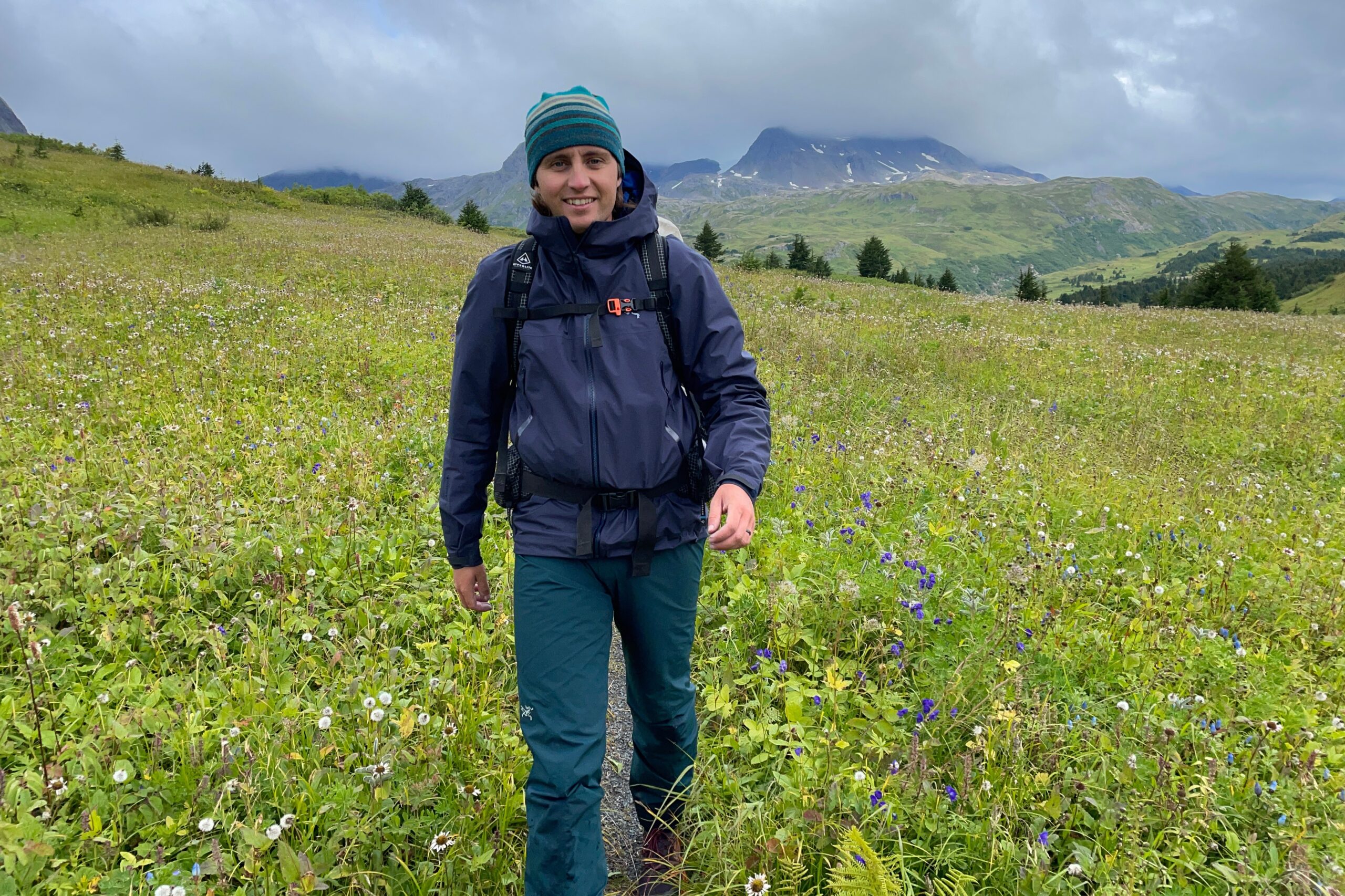 A man hikes through a field of wildflowers with mountains in the background