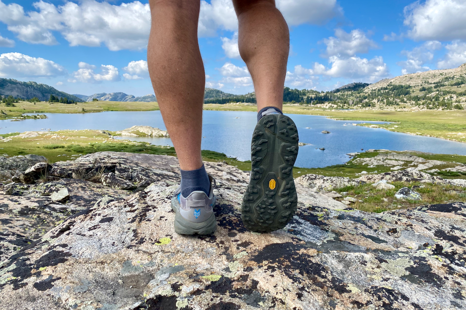 A hiker standing in front of a lake and mountains, looking away from the camera, showing the bottom of a pair of hiking shoes the HOKA Speedgoat 6.