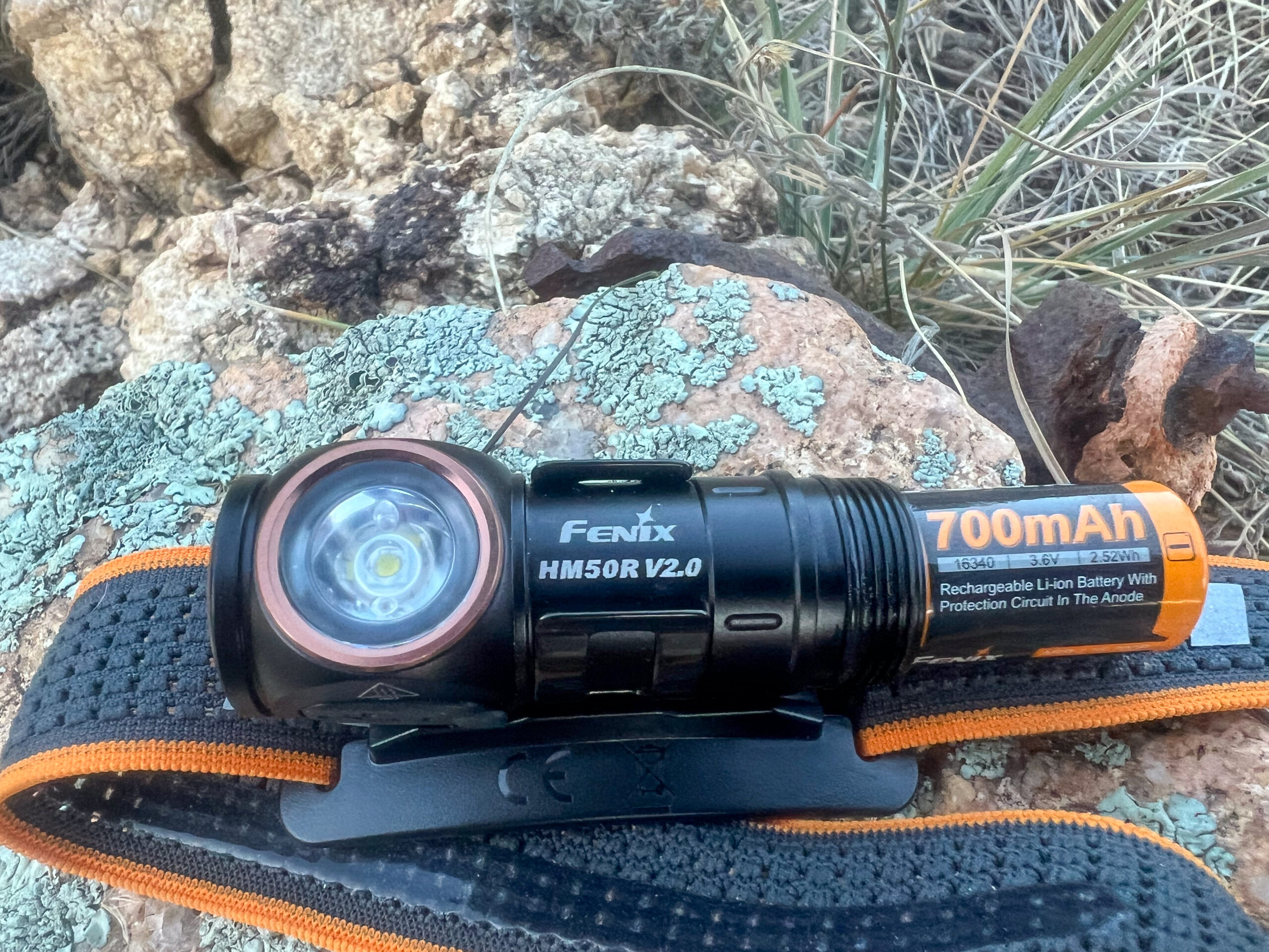 The image shows the Fenix HM50R V2.0 headlamp resting on a rock, with the rechargeable 700mAh battery clearly visible. The headlamp is attached to a black and orange headband with the Fenix logo, and the background consists of rocks and dried grass.