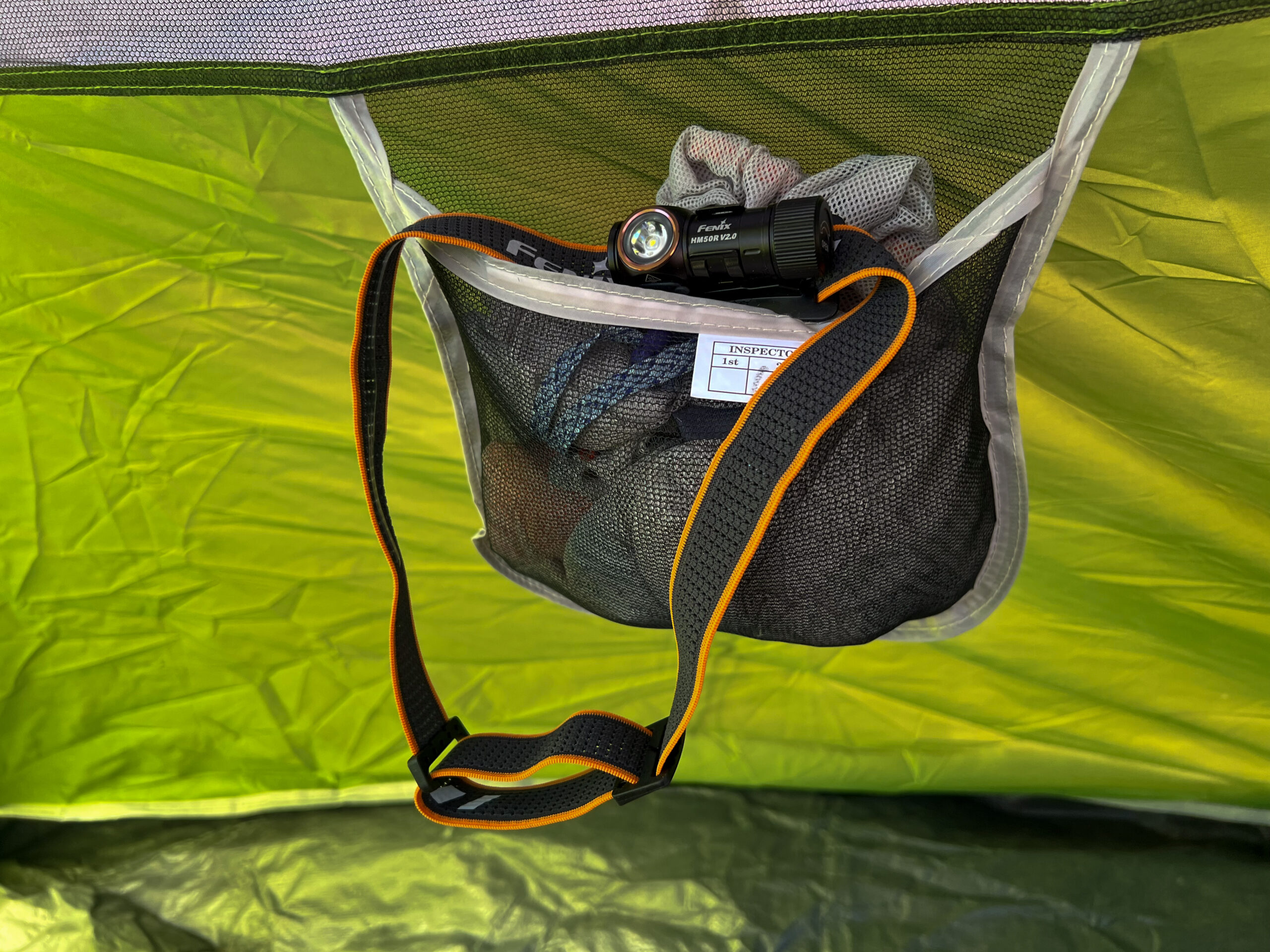 The image shows the Fenix HM50R V2.0 headlamp placed inside a mesh storage pocket within a tent. The headlamp is attached to its black and orange strap.