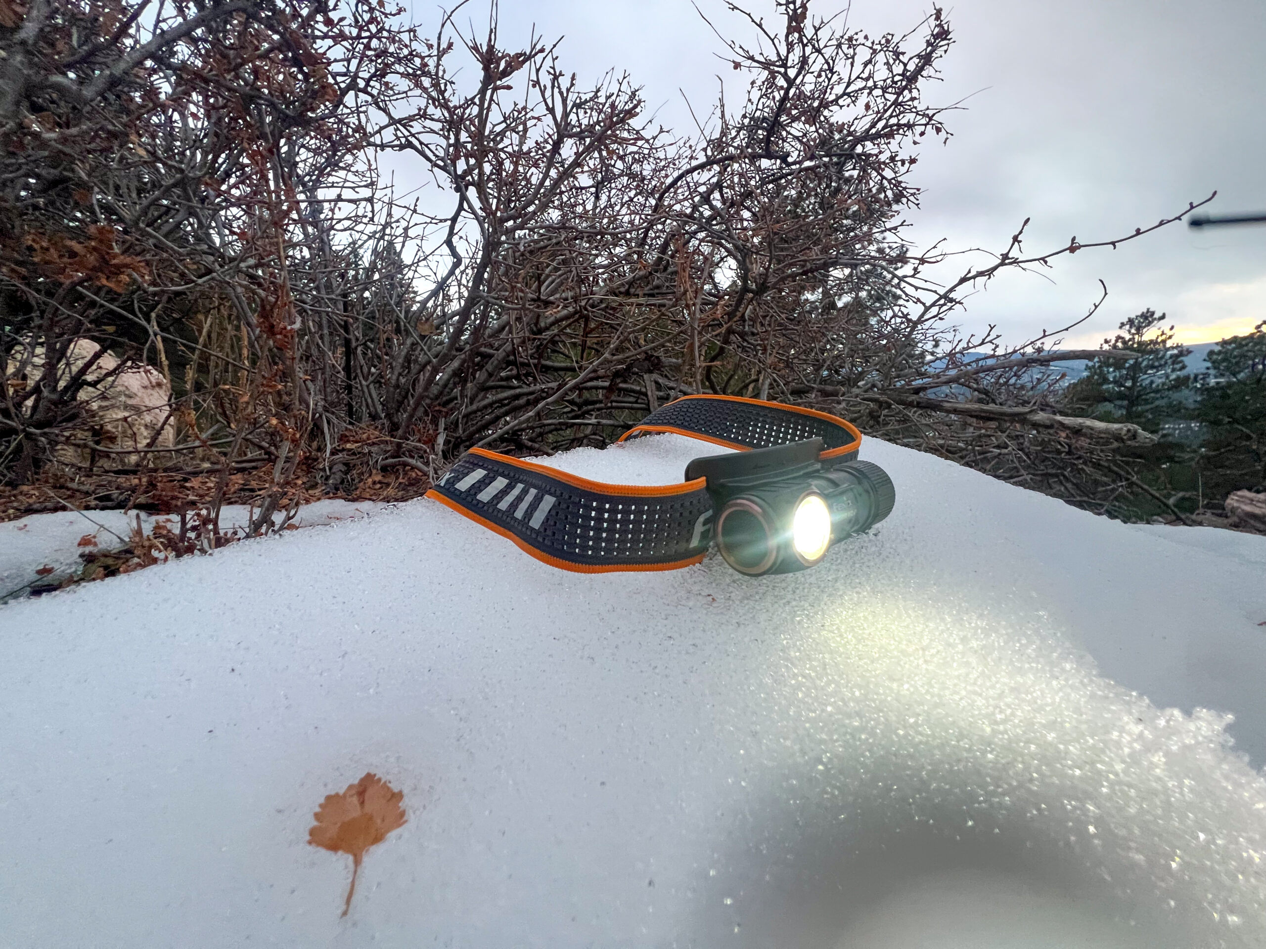 The image shows the Fenix HM50R V2.0 headlamp placed on snow, with the headlamp illuminated. The snow and surrounding area are slightly lit by the beam of the headlamp, which is casting a bright light.