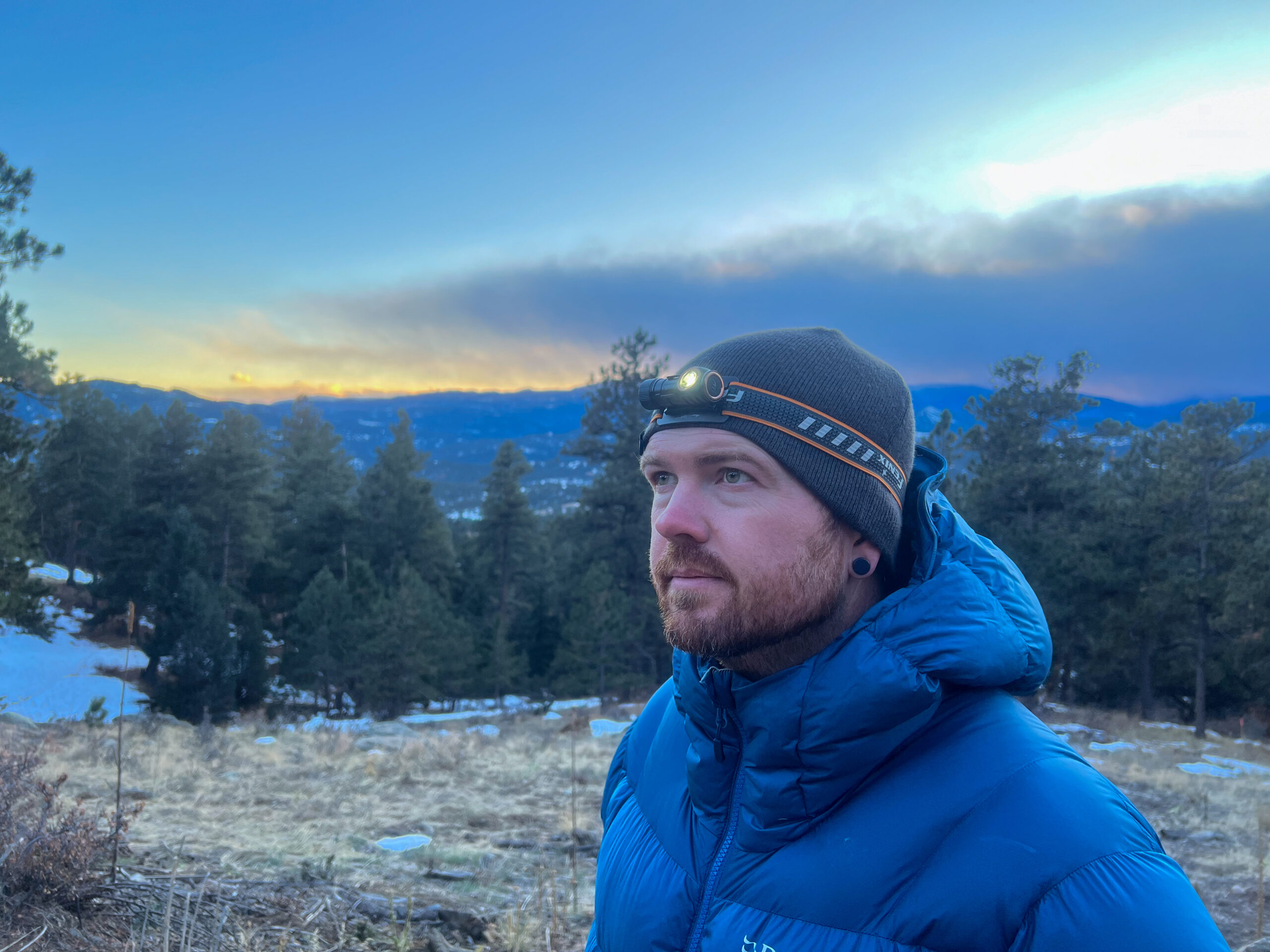 The image shows a person wearing the Fenix HM50R V2.0 headlamp while standing outdoors in a mountainous area. The person is looking towards the horizon, with a sunset visible in the background.