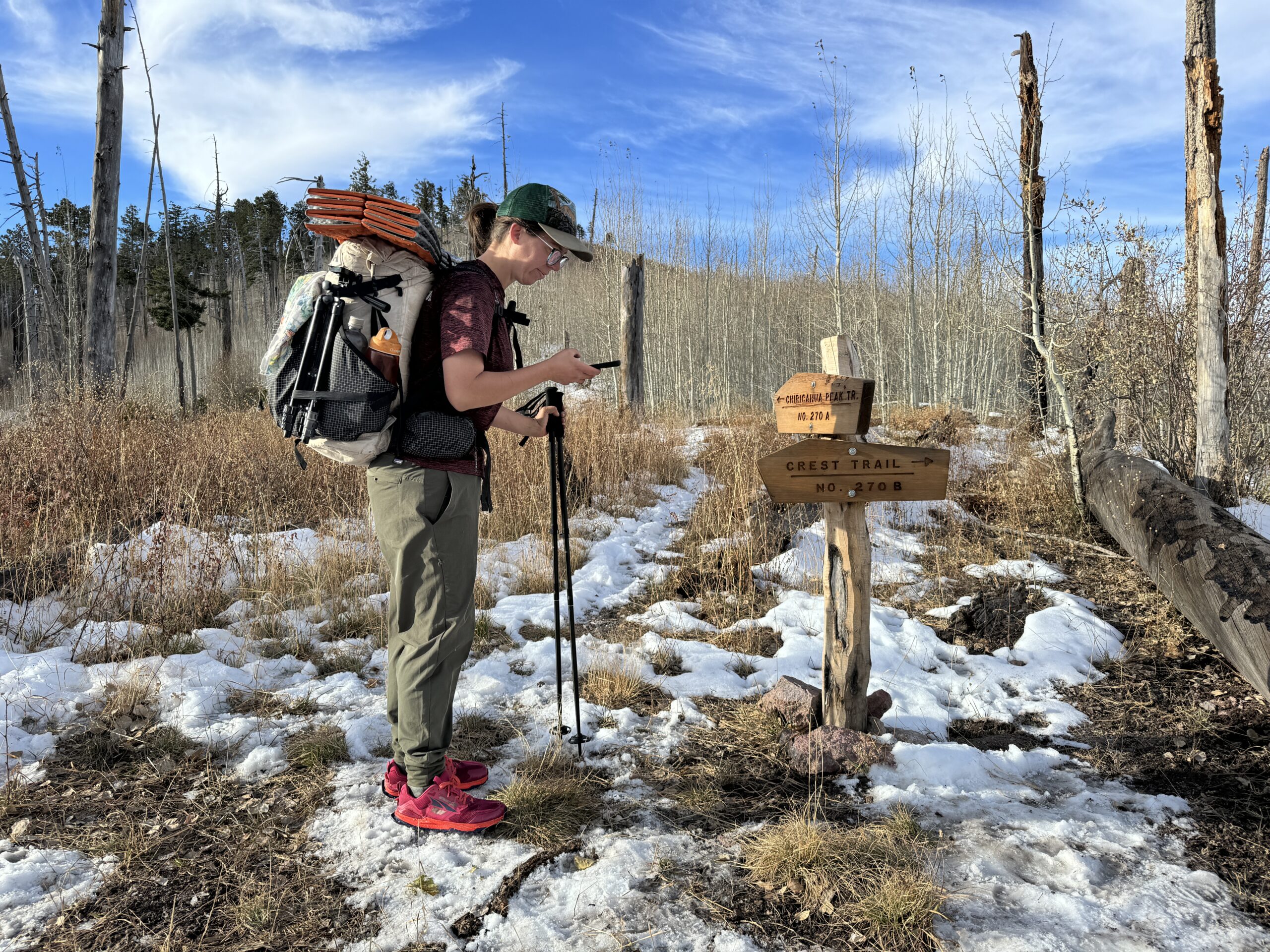 A hiker is looking at a phone next to a trail junction sign while wearing the Happy Hike pants in the woods.