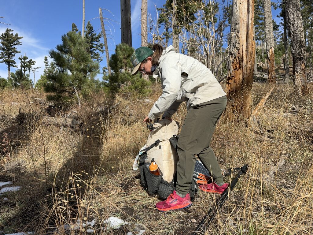 A hiker is packing up a backpack while wearing the Happy Hike pants in the woods.