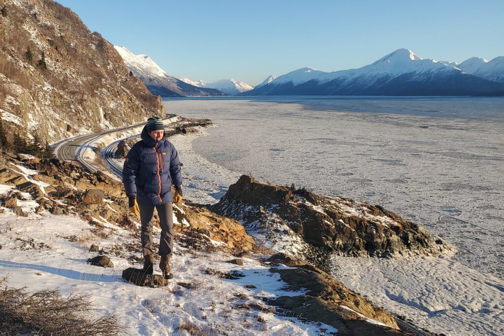 A man walks on a rocky outcropping above a mountain and ocean view.