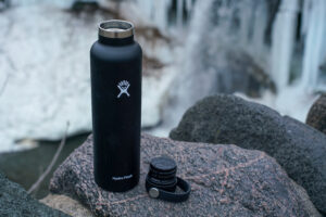 The Hydro Flask Standard Mouth, with its lid off, sits on a rock with a partially frozen waterfall in the background.