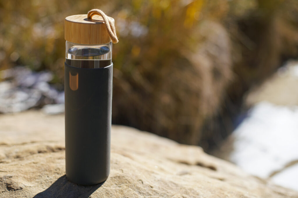 The Yomious Glass water bottle sits on a rock with a river in the background.