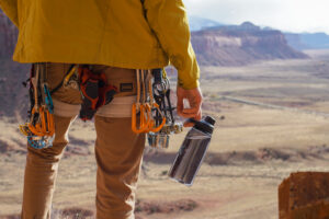 A person wearing a climbing harness holds the CamelBak Chute Mag Renew water bottle in their hand with a wide canyon expanse in the background.