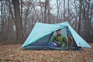 a man in a green coat sits in a one person tent in a forest clearing