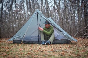 a man in a green coat puts his shoes on while sitting in a trekking pole tent