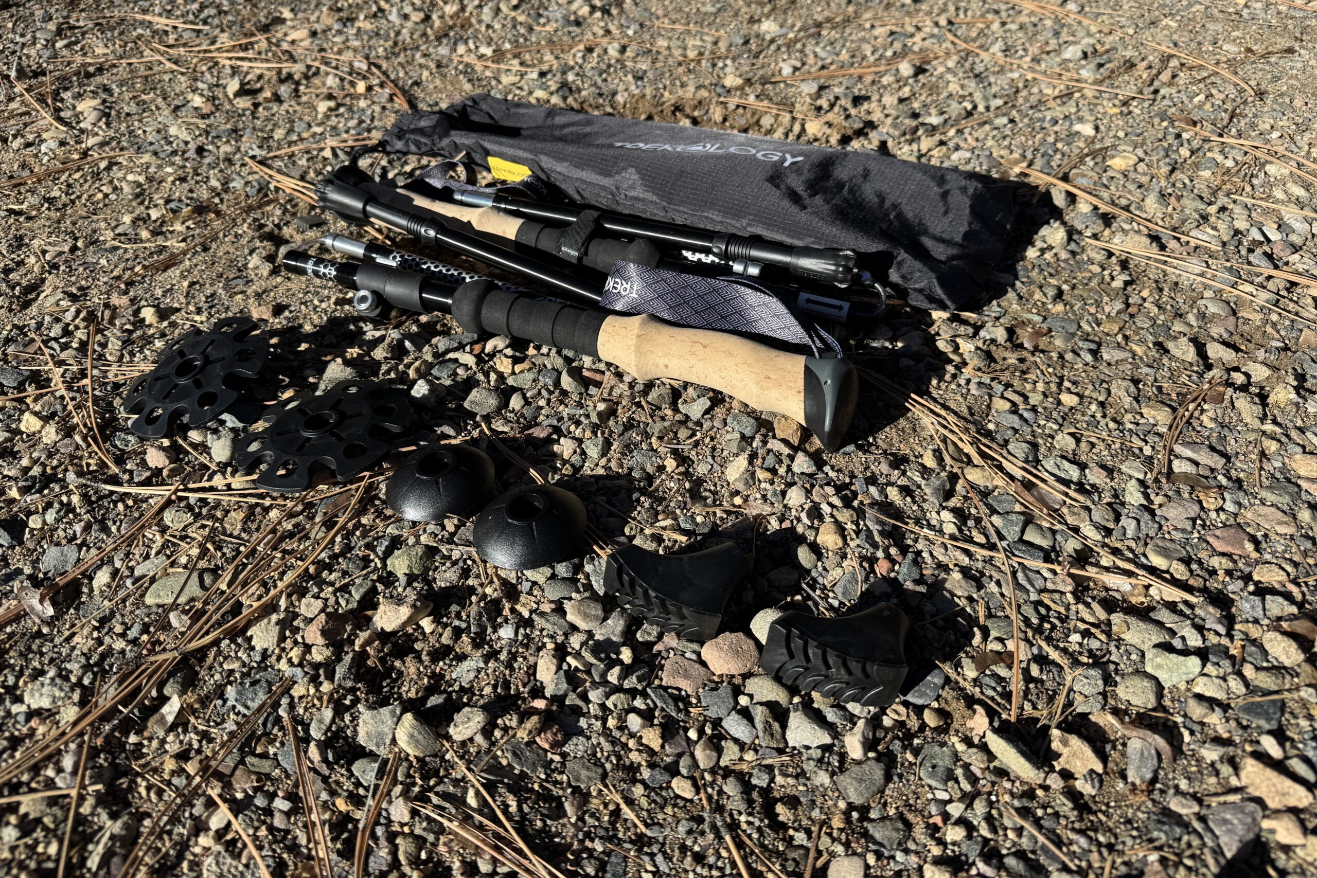 A collapsed trekking pole is laying on the ground in some gravel next to two baskets, four tip covers, and a storage bag.