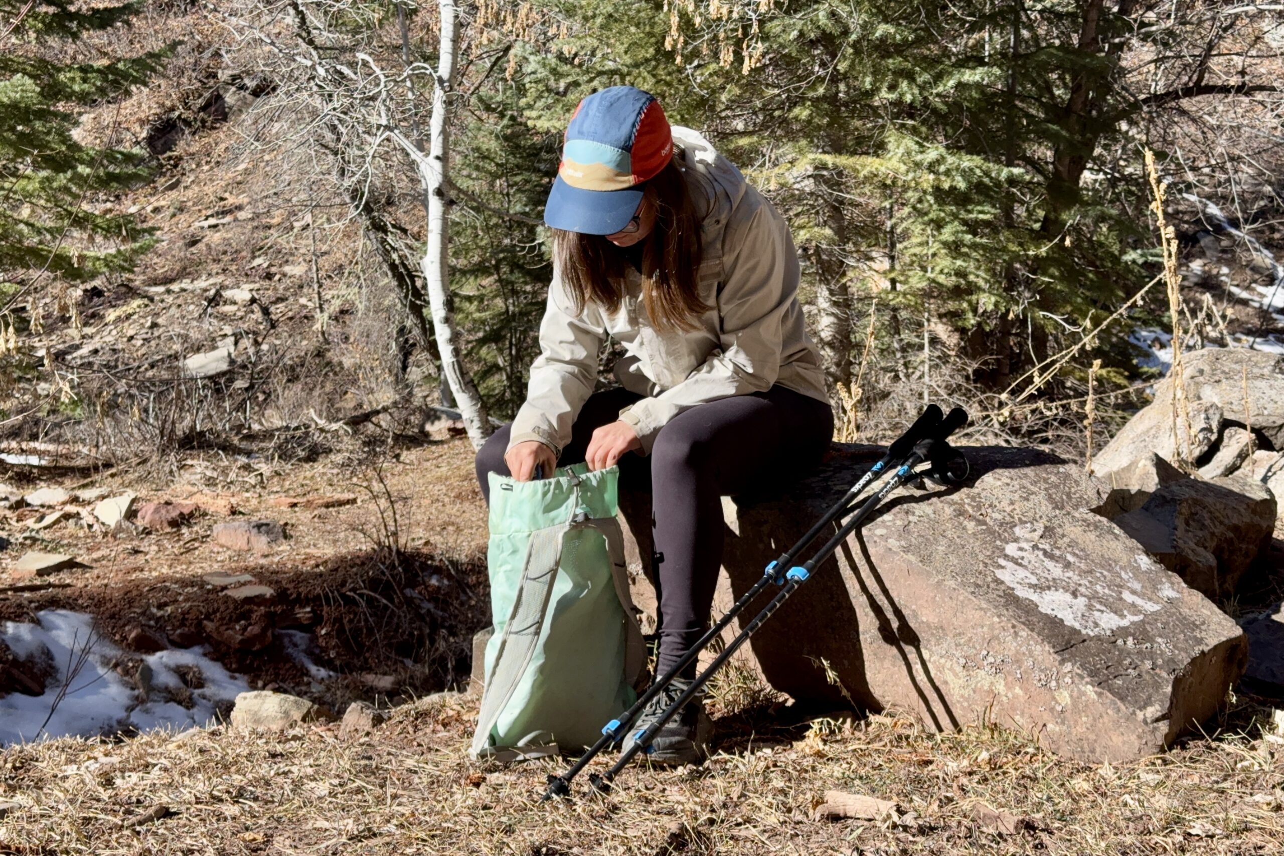 A person is sitting on a rock looking through a hiking bag. There is a pair of trekking poles leaning on the rock next to them. They are in a forest.