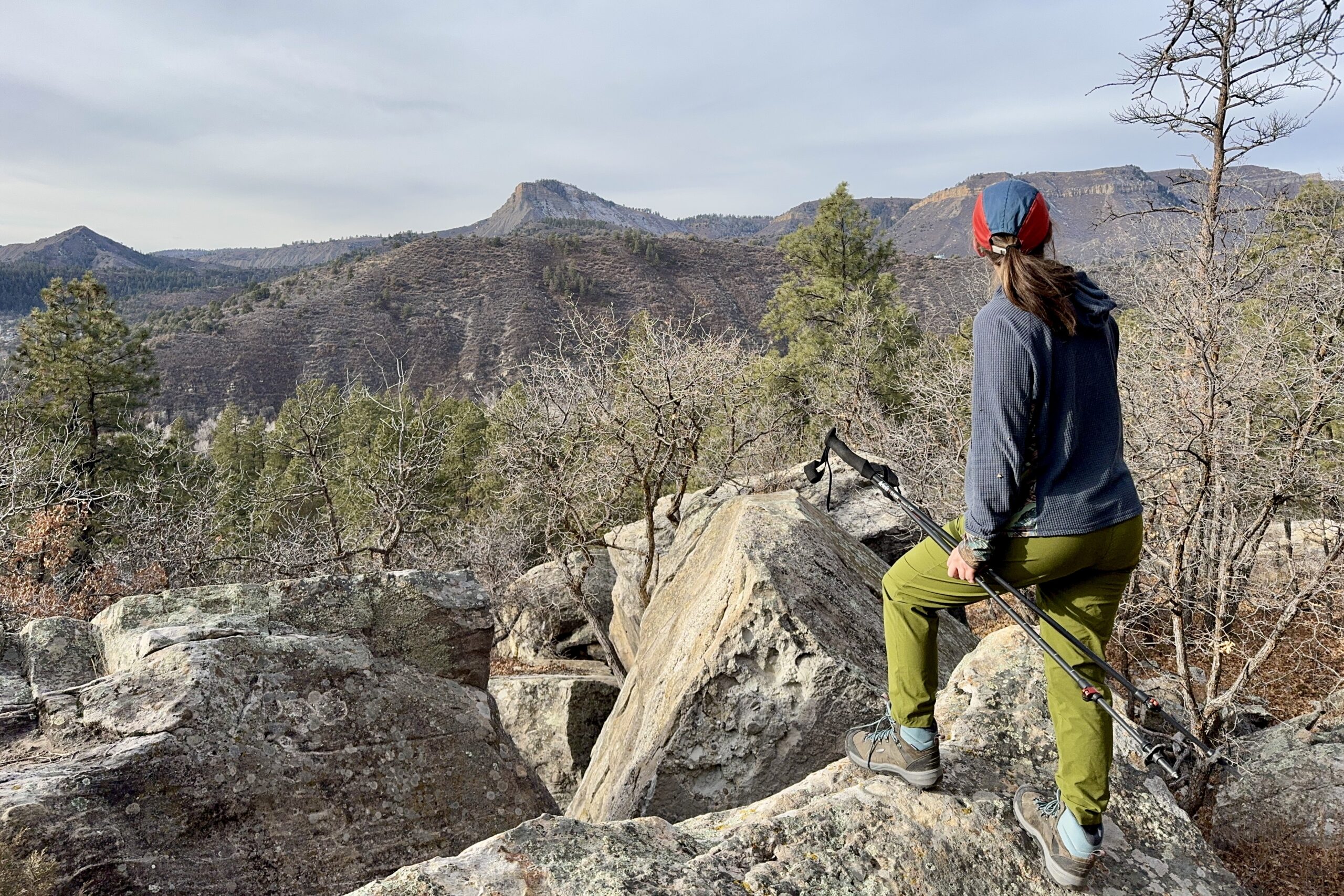 A person is standing on a rock and holding some trekking poles. They are looking out over a mountain view.