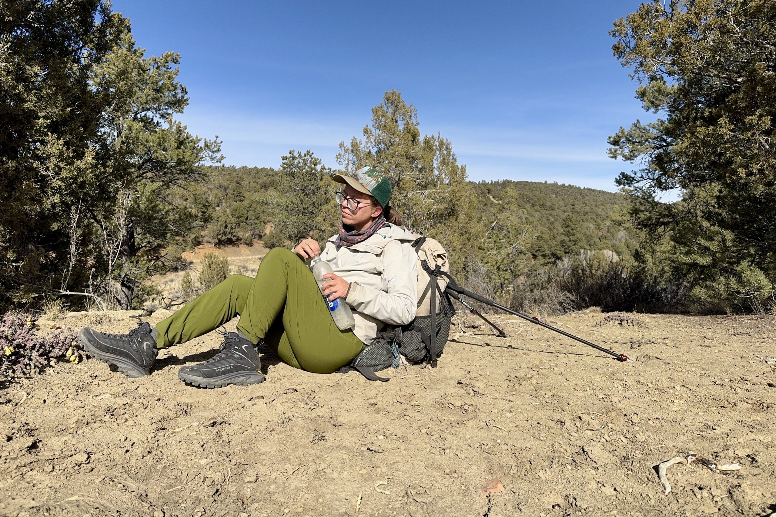 A person is relaxing by leaning on a backpack that is being held up by two trekking poles. The area is desert like with pine trees.