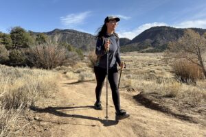 A person is walking down a trail using some trekking poles. They are in desert like area with bushes and pine trees.