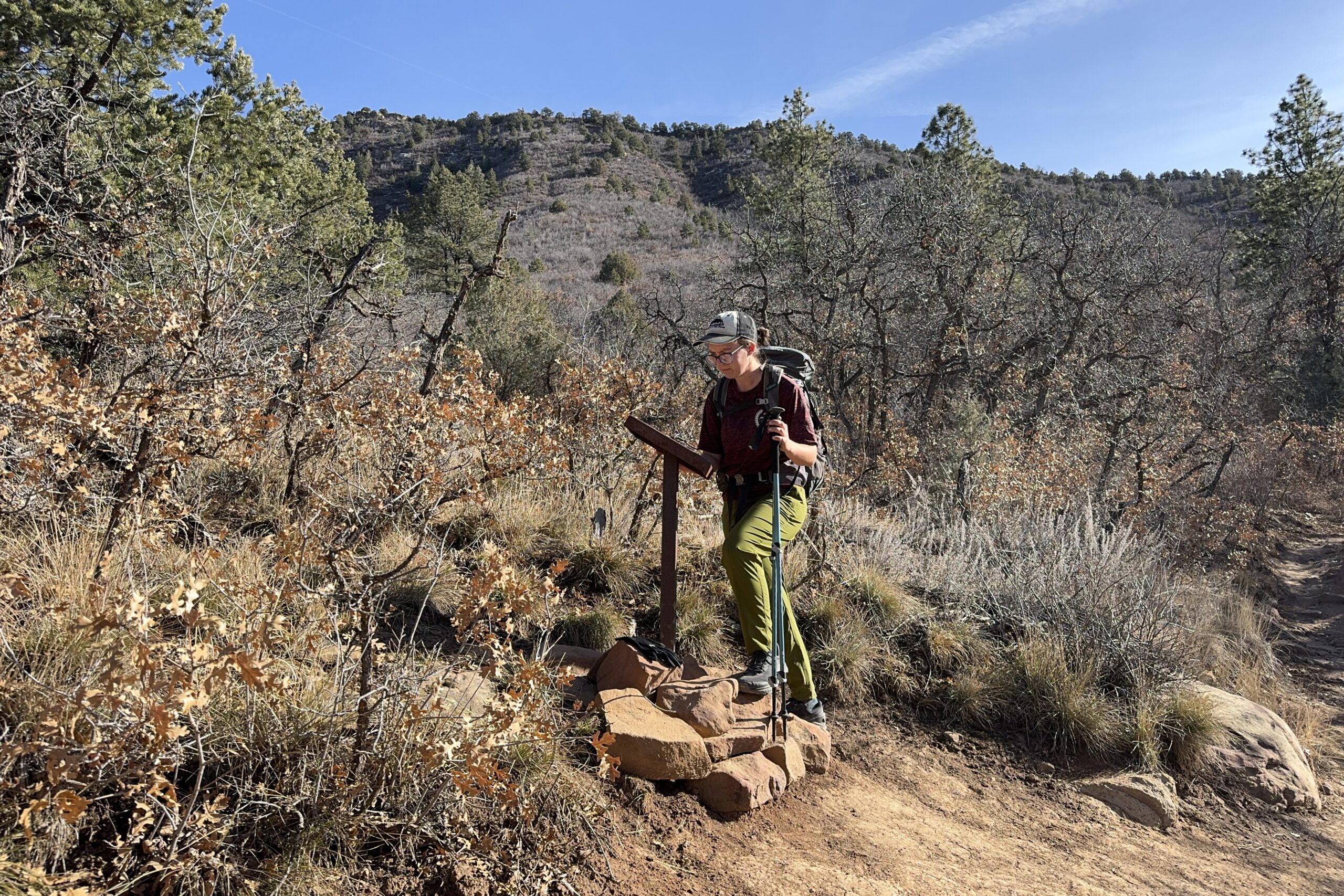 A person wearing a backpack is reading a trail map while holding some trekking poles. There are scrubby bushes in the background.