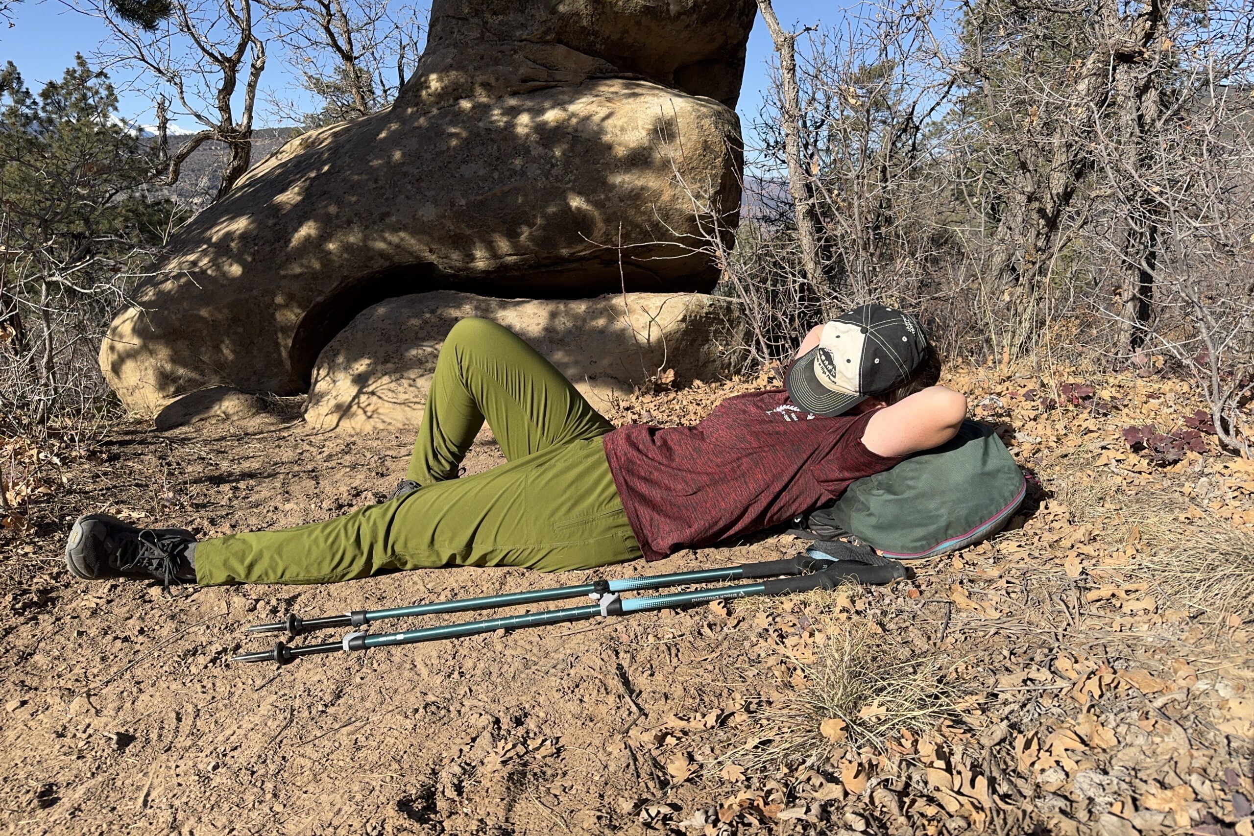 A person is laying on the ground using their backpack as a pillow with a hat over their face. There is a pair of trekking poles on the ground next to them and they are in a desert like area with trees and bushes.