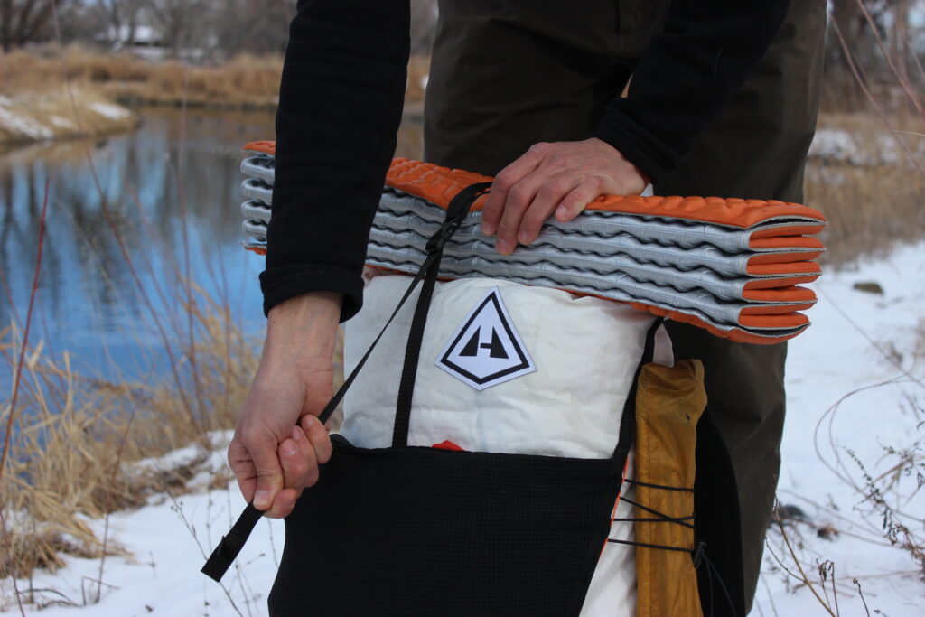 Close-up detail of the Hyperlite Mountain Gear Unbound backpack highlighting the roll-top closure system and its ability to secure a sleeping pad. The gray and orange accordion-style sleeping pad is mounted on top, secured by black compression straps. The scene is set against a winter backdrop with a partially frozen stream.