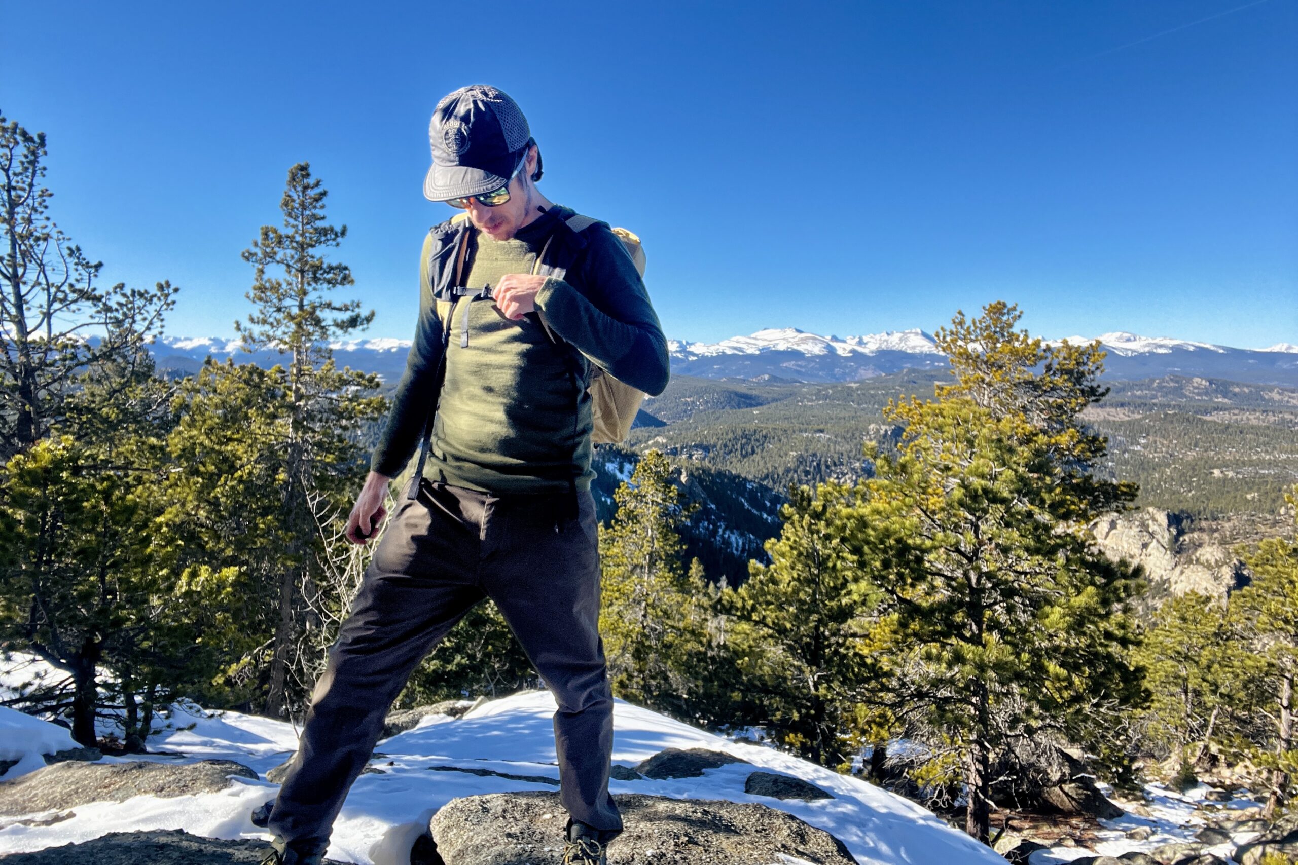 A hiker tightens the sternum strap on his backback while wearing the Smartwool Intraknit. Sky, mountains, and trees in the background.
