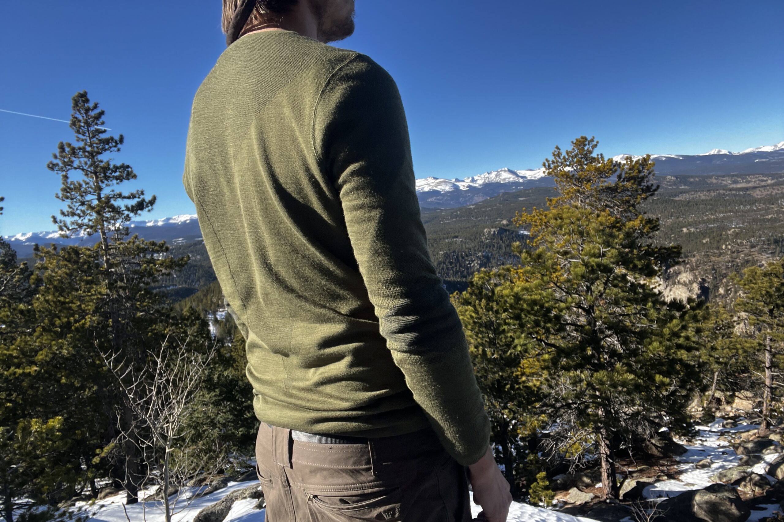 A hiker faces diagonally away from the camera wearing the Smartwool Intraknit base layer. Mountains, trees, and sky in the background.