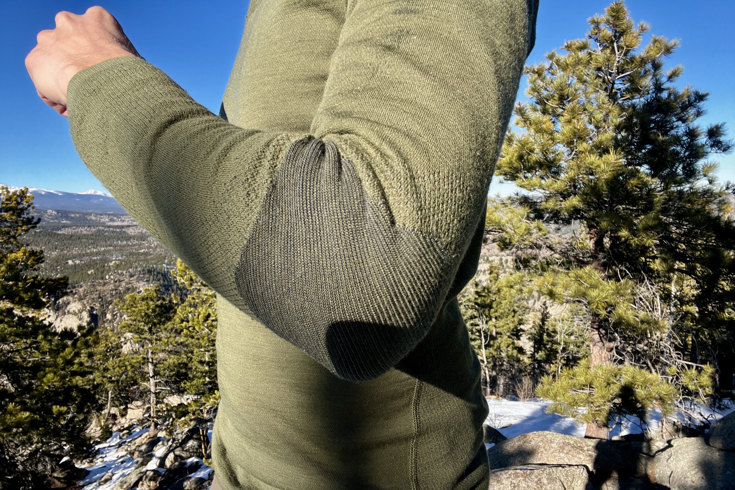 A hiker showing the elbow stitching of the Smartwool Intraknit. Trees and sky in the background.