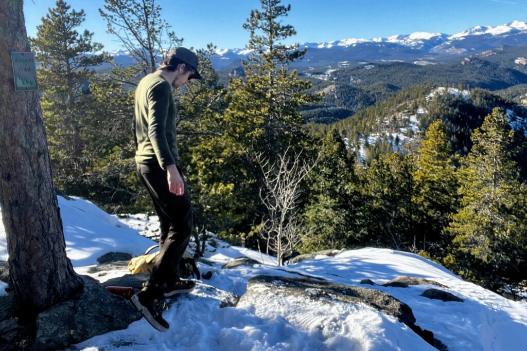 A hiker walks down snowy boulders with mountains in the background.