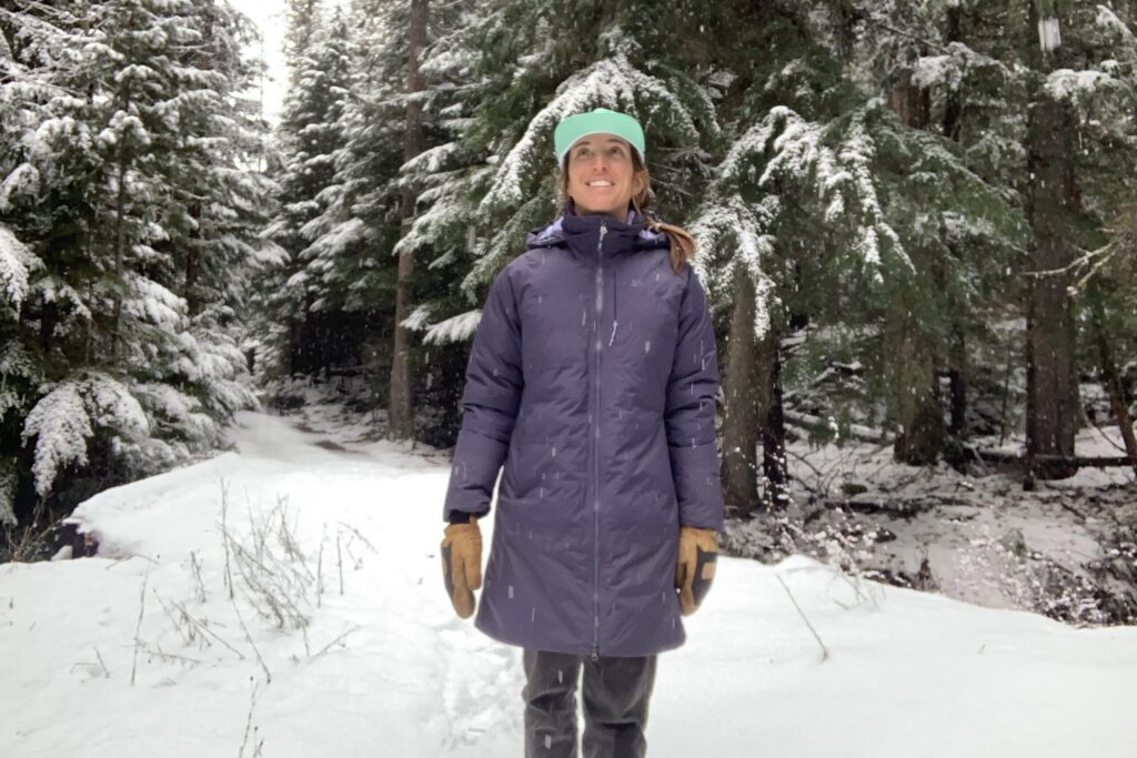 a woman standing in front of a snowy bridge in a forest looking at the snowfalling