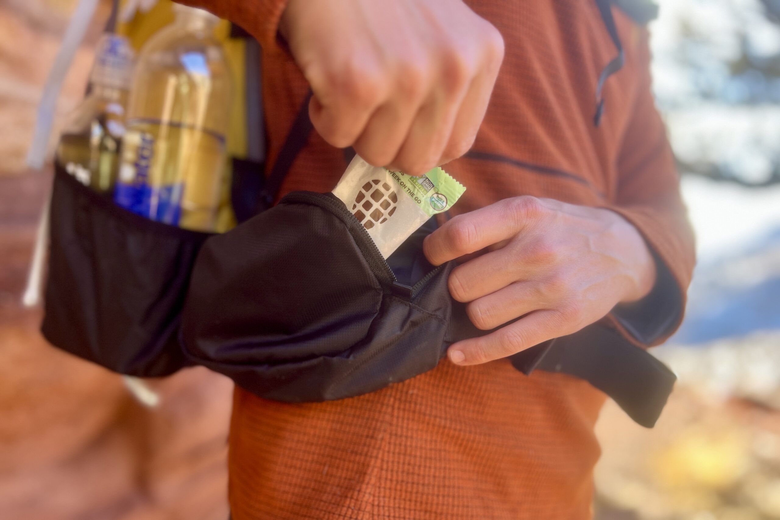 A person pulls a snack bar from the hip belt pocket of a backpack he is wearing.